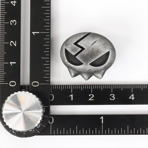 Peut inclure: Un charme en forme de cr&acirc;ne argent&eacute; avec un motif d'&eacute;clair et une surface textur&eacute;e. Le charme mesure environ 2,5 cm de diam&egrave;tre.