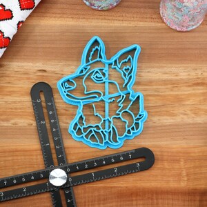May include: Blue plastic cookie cutter shaped like a dog's head. The cutter is on a wooden surface with a ruler and a spatula.