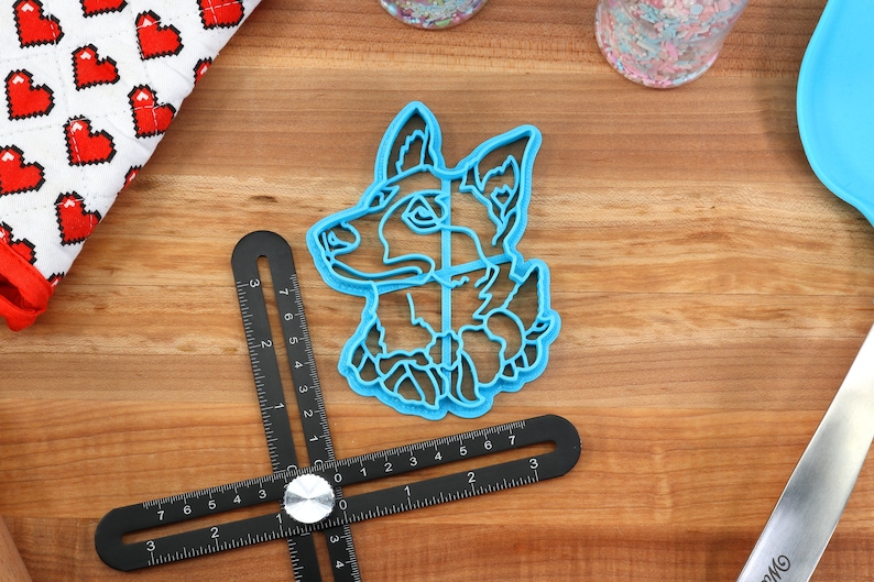 May include: Blue plastic cookie cutter shaped like a dog's head. The cutter is on a wooden surface with a ruler and a spatula.