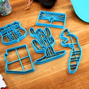 May include: A set of six blue plastic cookie cutters shaped like the state of Arizona, a cactus, a road sign that says "Arizona 66", a squirrel, and the Arizona state flag.