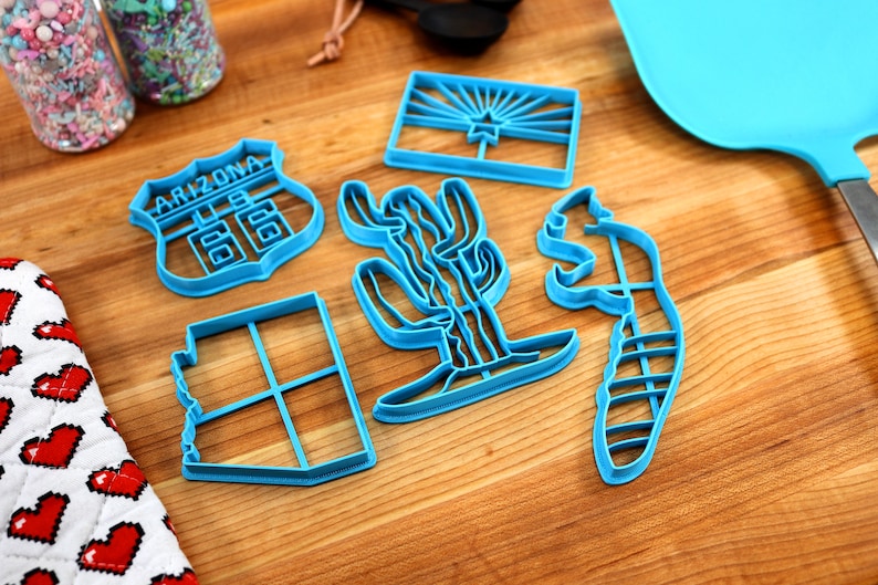 May include: A set of six blue plastic cookie cutters shaped like the state of Arizona, a cactus, a road sign that says "Arizona 66", a squirrel, and the Arizona state flag.