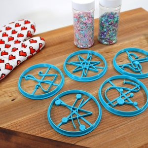 May include: Five blue plastic cookie cutters shaped like different scientific symbols on a wooden surface. Two jars of sprinkles are in the background.