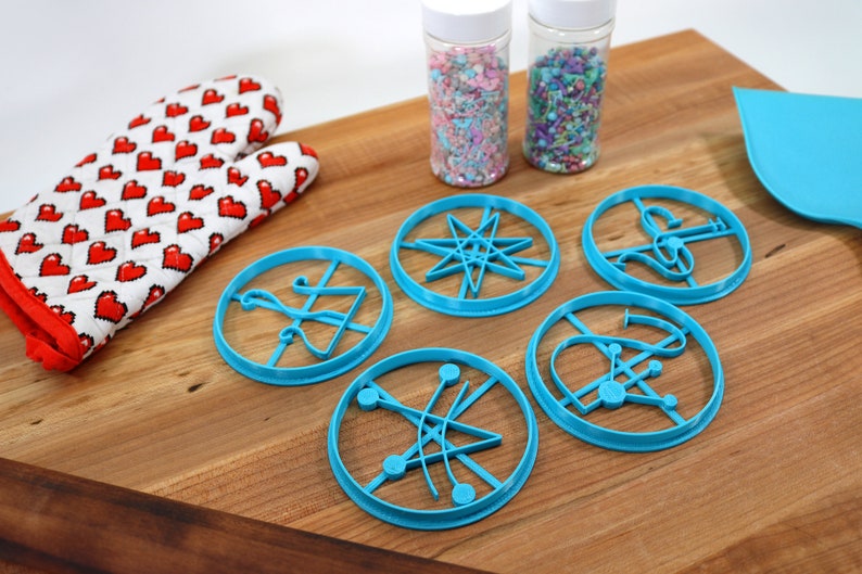 May include: Five blue plastic cookie cutters shaped like different scientific symbols on a wooden surface. Two jars of sprinkles are in the background.