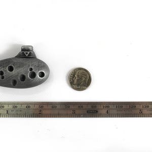 Puede incluir: Una ocarina plateada con un dise&ntilde;o triangular en la parte superior. La ocarina mide aproximadamente 5 cm de largo y est&aacute; sobre una superficie blanca junto a una regla y una moneda de diez centavos de d&oacute;lar estadounidense.