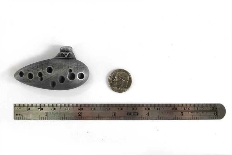 Puede incluir: Una ocarina plateada con un dise&ntilde;o triangular en la parte superior. La ocarina mide aproximadamente 5 cm de largo y est&aacute; sobre una superficie blanca junto a una regla y una moneda de diez centavos de d&oacute;lar estadounidense.
