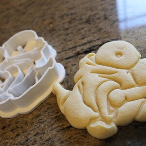 May include: A white plastic cookie cutter next to a baked sugar cookie. The cookie cutter is in the shape of a character. The baked cookie is light brown and has detailed impressions from the cutter.