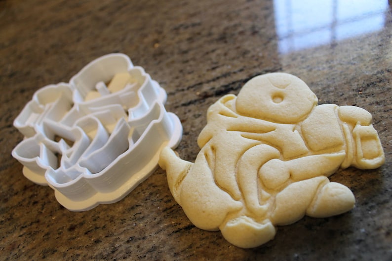 May include: A white plastic cookie cutter next to a baked sugar cookie. The cookie cutter is in the shape of a character. The baked cookie is light brown and has detailed impressions from the cutter.