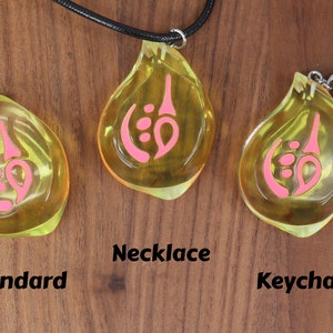 May include: Three yellow teardrop-shaped pendants with pink designs. The pendant on the left is labeled "Standard", the middle pendant is on a black cord and labeled "Necklace", and the pendant on the right is attached to a silver keychain and labeled "Keychain".