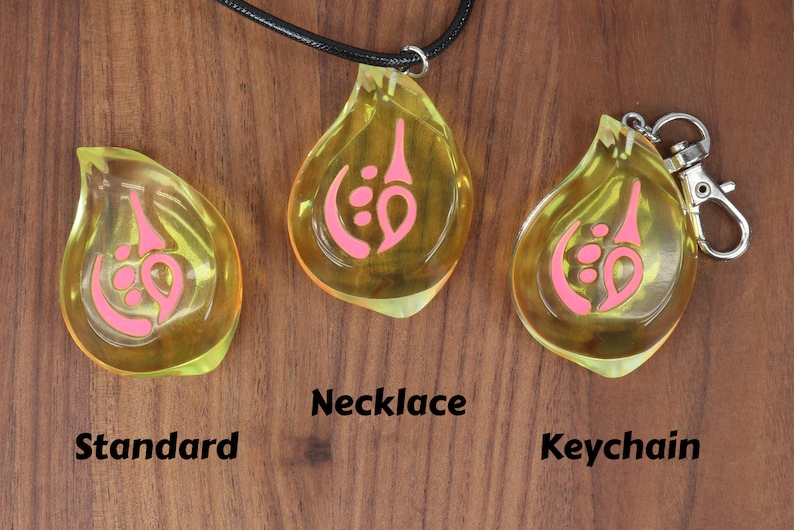 May include: Three yellow teardrop-shaped pendants with pink designs. The pendant on the left is labeled "Standard", the middle pendant is on a black cord and labeled "Necklace", and the pendant on the right is attached to a silver keychain and labeled "Keychain".