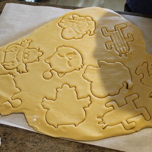 May include: A sheet of raw cookie dough with various cut-out shapes, including birds and other whimsical figures. The dough is light brown, and the cut-out shapes are clearly defined. The scene suggests a baking activity.