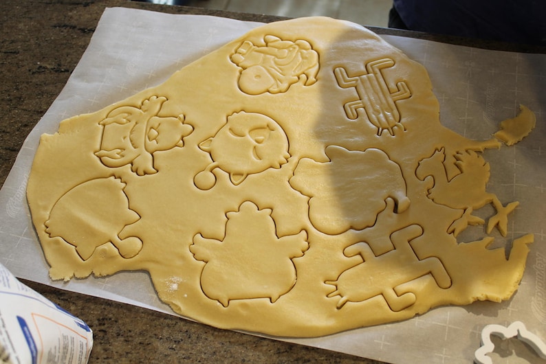 May include: A sheet of raw cookie dough with various cut-out shapes, including birds and other whimsical figures. The dough is light brown, and the cut-out shapes are clearly defined. The scene suggests a baking activity.