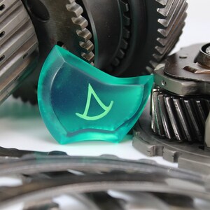 May include: A close-up of a green plastic gear with a white sailboat symbol on it. The gear is surrounded by metal gears.