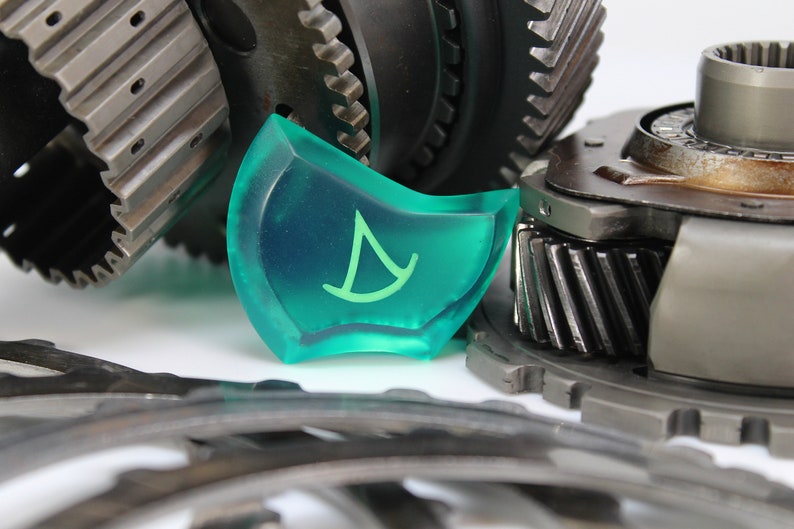 May include: A close-up of a green plastic gear with a white sailboat symbol on it. The gear is surrounded by metal gears.