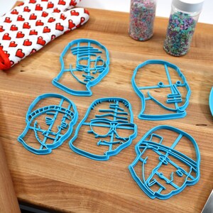 May include: Five blue plastic cookie cutters shaped like different faces. The cutters are on a wooden surface with a rolling pin, a spatula, and two jars of sprinkles.