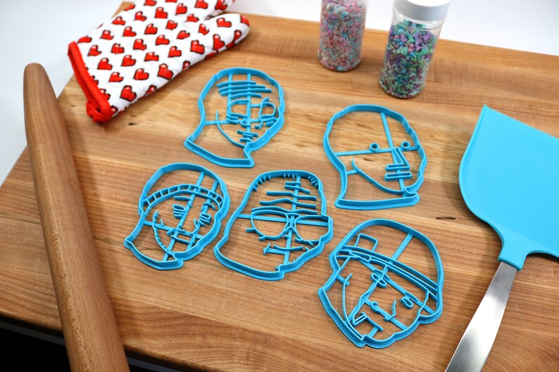 May include: Five blue plastic cookie cutters shaped like different faces. The cutters are on a wooden surface with a rolling pin, a spatula, and two jars of sprinkles.