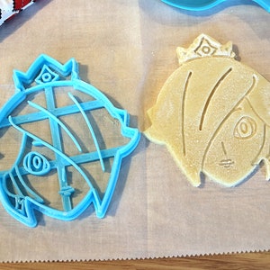 May include: A blue plastic cookie cutter shaped like a cartoon character with a crown, next to a baked cookie cut with the same shape.