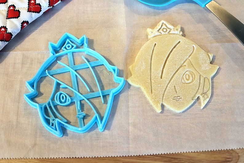 May include: A blue plastic cookie cutter shaped like a cartoon character with a crown, next to a baked cookie cut with the same shape.
