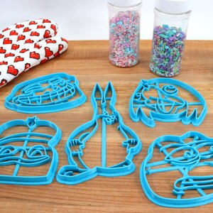 Puede incluir: Conjunto de seis cortadores de galletas de pl&aacute;stico azul con forma de personajes de dibujos animados. Los cortadores est&aacute;n sobre una superficie de madera con dos frascos de chispas.