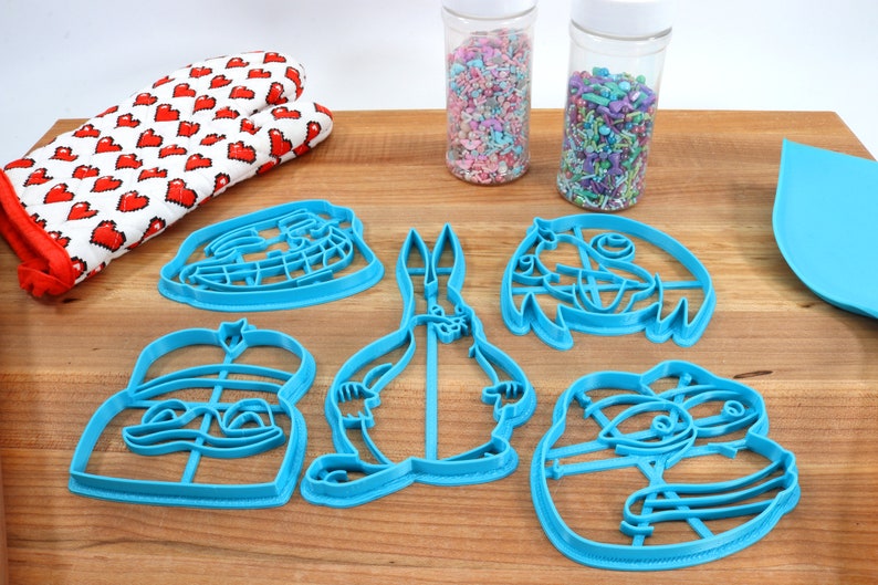 Puede incluir: Conjunto de seis cortadores de galletas de pl&aacute;stico azul con forma de personajes de dibujos animados. Los cortadores est&aacute;n sobre una superficie de madera con dos frascos de chispas.