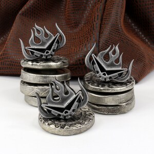 May include: Three silver metal dice with a skull and flame design, stacked on top of each other. The dice are sitting on a white surface.