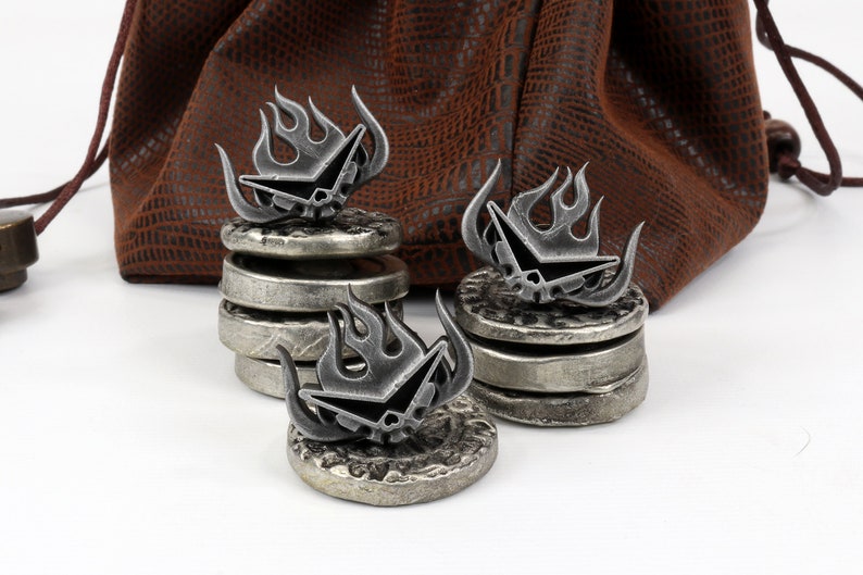 May include: Three silver metal dice with a skull and flame design, stacked on top of each other. The dice are sitting on a white surface.