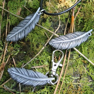 Op de afbeelding: Een verzameling zilverkleurige veervormige hangers en bedels. E&eacute;n veer hangt aan een zwarte koordketting, twee zijn los en &eacute;&eacute;n is bevestigd aan een zilverkleurige sluiting. De items liggen op mos en twijgen.