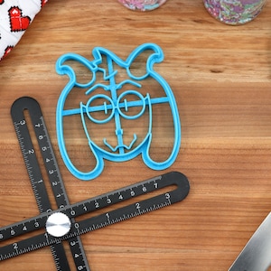 Peut inclure: Un emporte-pi&egrave;ce en plastique bleu clair en forme de visage de chien de dessin anim&eacute;. Un outil de mesure r&eacute;glable noir et une spatule bleue sont &eacute;galement visibles sur une surface en bois. Un gant de cuisine &agrave; motifs de c&oelig;urs rouges et blancs est en arri&egrave;re-plan.