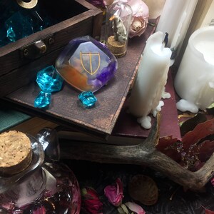 May include: A wooden box filled with blue glass gems, a clear resin pendant with a gold and purple design, a small glass vial with a dried flower inside, a white candle, and a wooden antler.