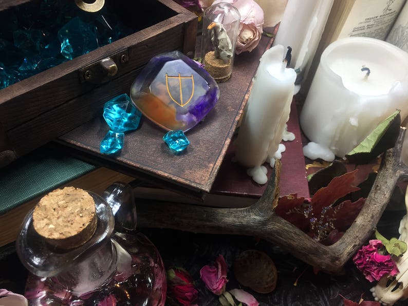 May include: A wooden box filled with blue glass gems, a clear resin pendant with a gold and purple design, a small glass vial with a dried flower inside, a white candle, and a wooden antler.