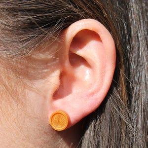 May include: A close-up of a person's ear with a small, round, gold-colored earring. The earring has a raised design.