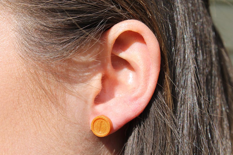 May include: A close-up of a person's ear with a small, round, gold-colored earring. The earring has a raised design.