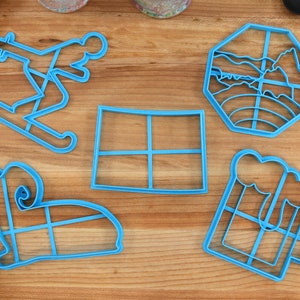 Puede incluir: Cinco cortadores de galletas de plástico azul sobre una superficie de madera. Los cortadores tienen forma de persona esquiando, una ventana, una cordillera, una jarra de cerveza y un perro.