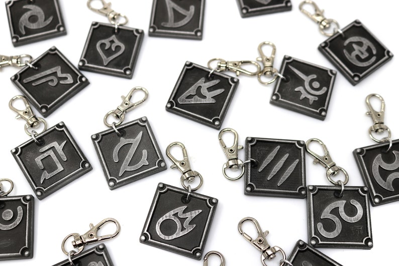May include: A set of 15 silver and black metal keychains with different symbols. Each keychain features a square-shaped charm with a unique symbol in a silver color on a black background.