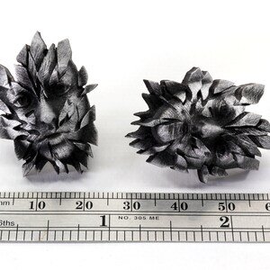 Pu&ograve; includere: Due sculture in metallo color argento di volti, ciascuna con un design geometrico frammentato. Le sculture hanno un diametro di circa 5 cm e sono posizionate su una superficie bianca con un righello per la scala.