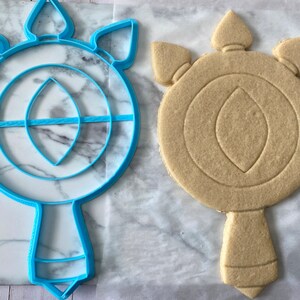 May include: A blue plastic cookie cutter shaped like a wand with a circular top and a pointed bottom. Next to it is a light brown sugar cookie that has been cut out using the cookie cutter.