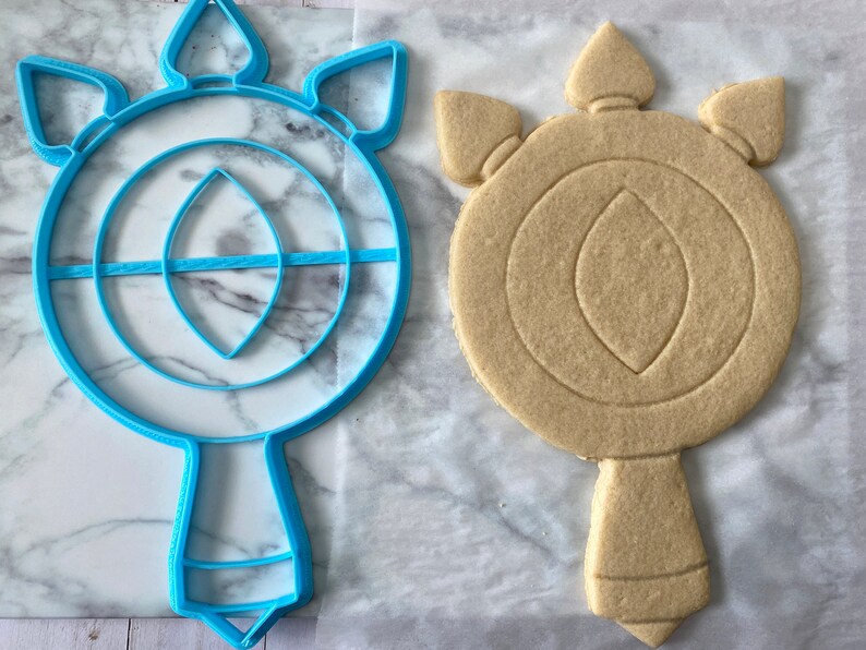 May include: A blue plastic cookie cutter shaped like a wand with a circular top and a pointed bottom. Next to it is a light brown sugar cookie that has been cut out using the cookie cutter.