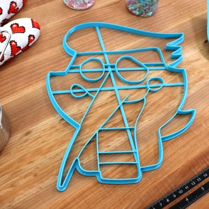 May include: A blue plastic cookie cutter shaped like a pirate with a hat, eye patch, and sword. The cutter is on a wooden surface.