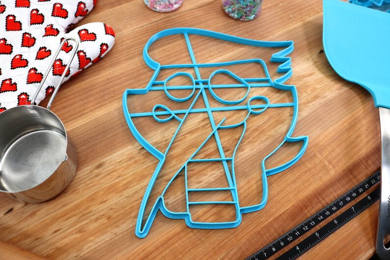 May include: A blue plastic cookie cutter shaped like a pirate with a hat, eye patch, and sword. The cutter is on a wooden surface.