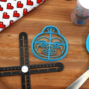 May include: A blue plastic sloth-shaped cookie cutter with a smiling face and a small hat on top. The cutter is on a wooden surface with a ruler and a rolling pin.