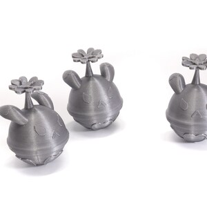 May include: Three gray 3D printed figurines of a cartoon character with a flower on top. The character has a round body, two ears, and a small nose.