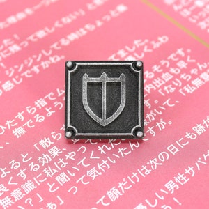 Puede incluir: Un pin de metal de color plateado con un dise&ntilde;o cuadrado. El pin presenta un escudo con tres puntas en la parte superior y un escudo m&aacute;s peque&ntilde;o en el interior. El pin est&aacute; sobre un fondo rosa con texto japon&eacute;s.