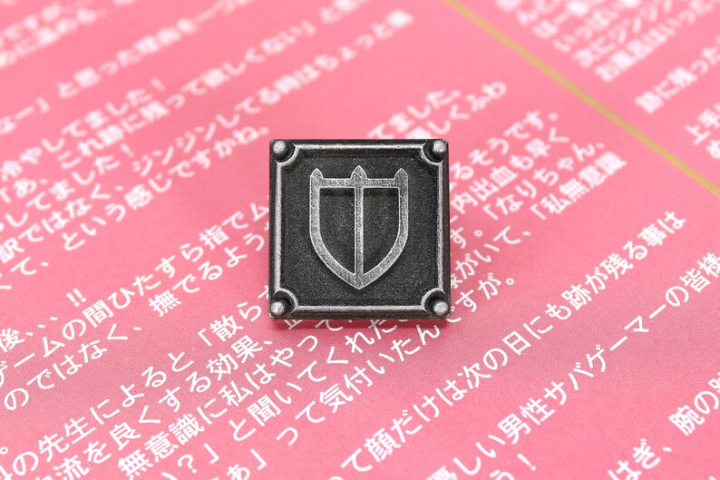 Puede incluir: Un pin de metal de color plateado con un dise&ntilde;o cuadrado. El pin presenta un escudo con tres puntas en la parte superior y un escudo m&aacute;s peque&ntilde;o en el interior. El pin est&aacute; sobre un fondo rosa con texto japon&eacute;s.