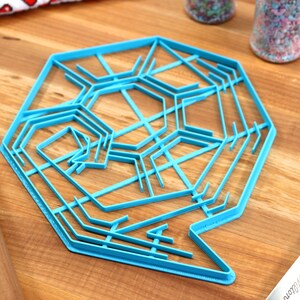 May include: A blue plastic snowflake cookie cutter with a detailed design. The cutter is on a wooden surface.