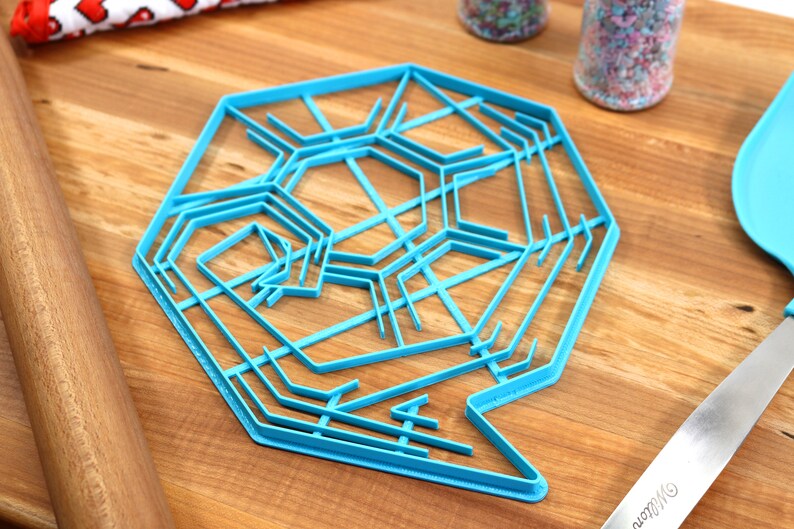 May include: A blue plastic snowflake cookie cutter with a detailed design. The cutter is on a wooden surface.