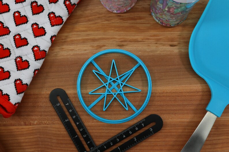 May include: A blue plastic cookie cutter shaped like a star with eight points. The cutter is on a wooden surface with a ruler and a blue silicone spatula.