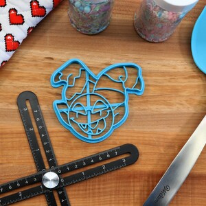 May include: A blue plastic cookie cutter shaped like an elf's face with a mischievous grin. The cutter is on a wooden surface with a ruler and a spatula.