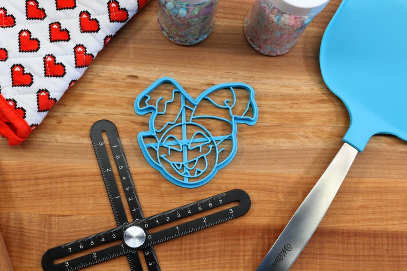 May include: A blue plastic cookie cutter shaped like an elf's face with a mischievous grin. The cutter is on a wooden surface with a ruler and a spatula.
