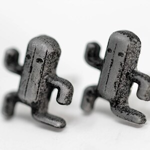May include: A pair of silver-colored earrings shaped like a video game character. The earrings are detailed with a textured surface and have a slightly worn look.