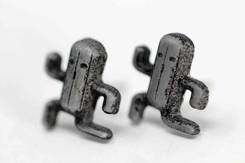 May include: A pair of silver-colored earrings shaped like a video game character. The earrings are detailed with a textured surface and have a slightly worn look.