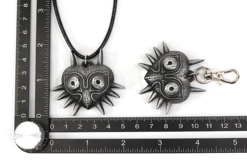 May include: Two silver Majora's Mask pendants, one on a black cord necklace and the other on a silver key chain. The pendants are both heart-shaped with a detailed design of the Majora's Mask from the Legend of Zelda video game.